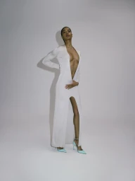 Model Raven Wallace posing in a plunging white long-sleeve slit gown and light blue pointed heels against a white studio backdrop.