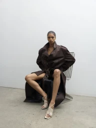 Model Raven Wallace seated in a wire chair wearing an oversized dark brown trench coat and cream woven heeled sandals.