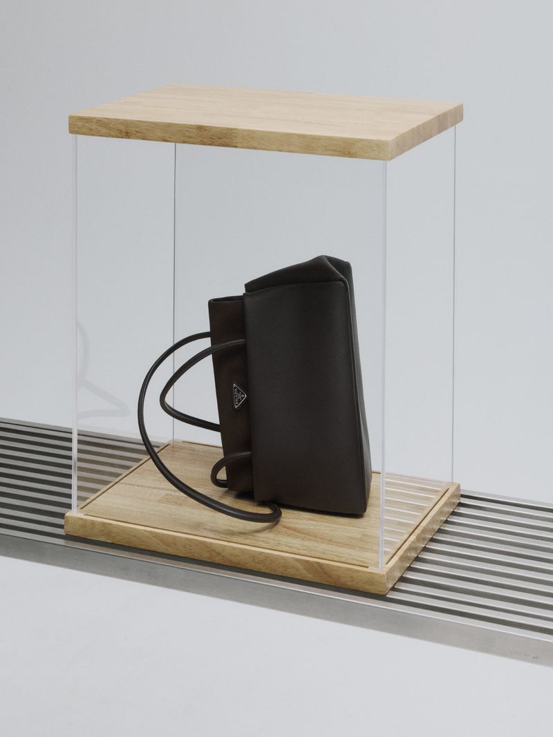 Dark brown leather handbag displayed inside a clear acrylic and wood vitrine resting on a slatted metal surface.