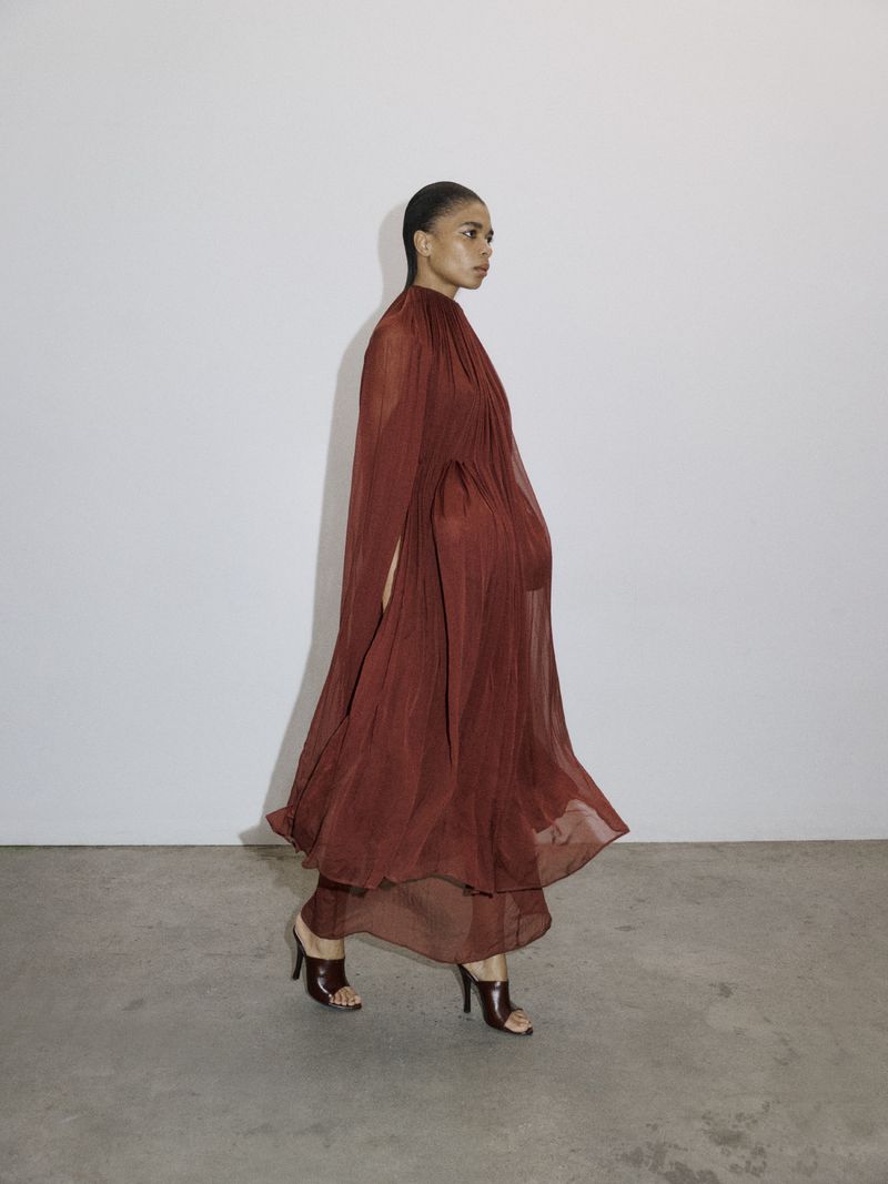 Model Raven Wallace walking in a sheer pleated terracotta dress and dark brown heeled mules against a studio backdrop.