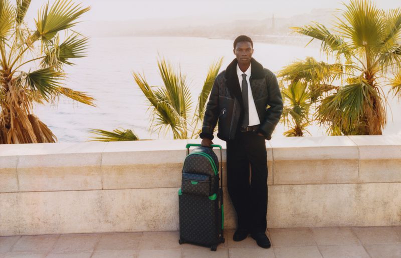A dark-skinned man stands outside near palm trees with a travel suitcase and a backpack, dressed in a jacket, white shirt, and black pants.
