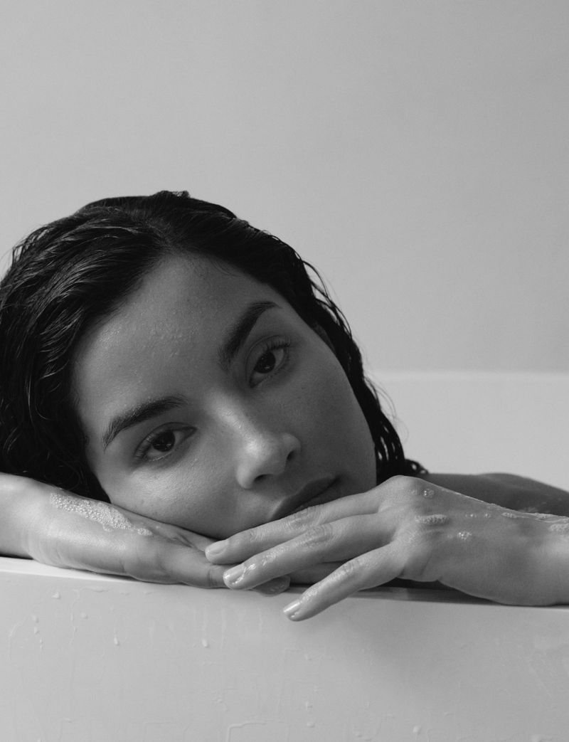 Monochrome close-up of model Tindi Mar resting her head and wet hands on the ledge of a white bathtub.