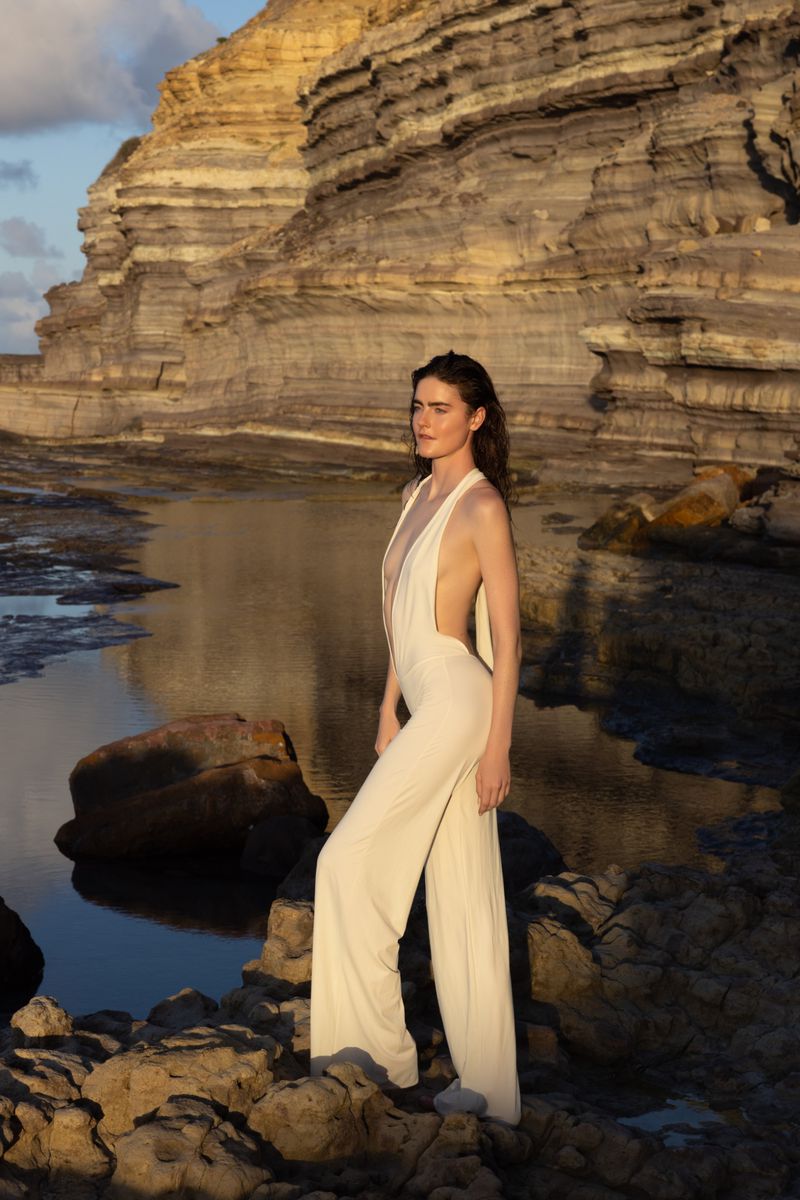 Daphne Velghe in a cream backless jumpsuit standing on a rocky shoreline beneath coastal cliffs.