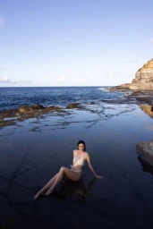 Model Daphne Velghe reclining in a shallow rock pool wearing a cream swimsuit along a rocky ocean coastline.