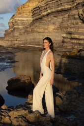 Daphne Velghe in a cream backless jumpsuit standing on a rocky shoreline beneath coastal cliffs.