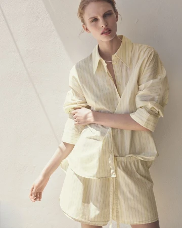 Model Karolina Spakowski wearing a pale yellow striped button-up shirt and matching shorts in bright sunlight against a wall.