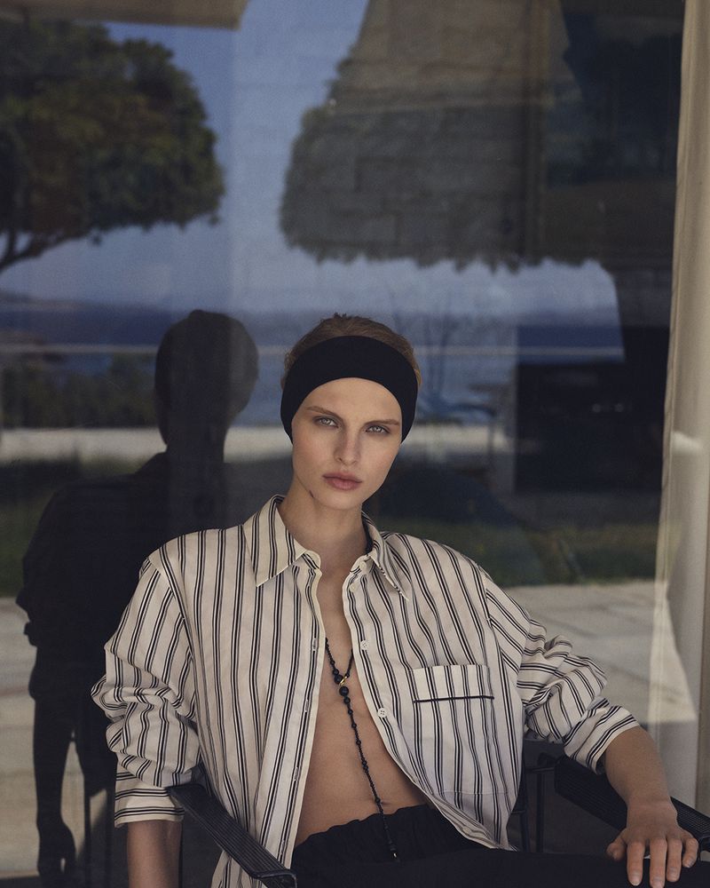 Model Karolina Spakowski seated in an unbuttoned striped shirt and black headband against a reflective glass window.