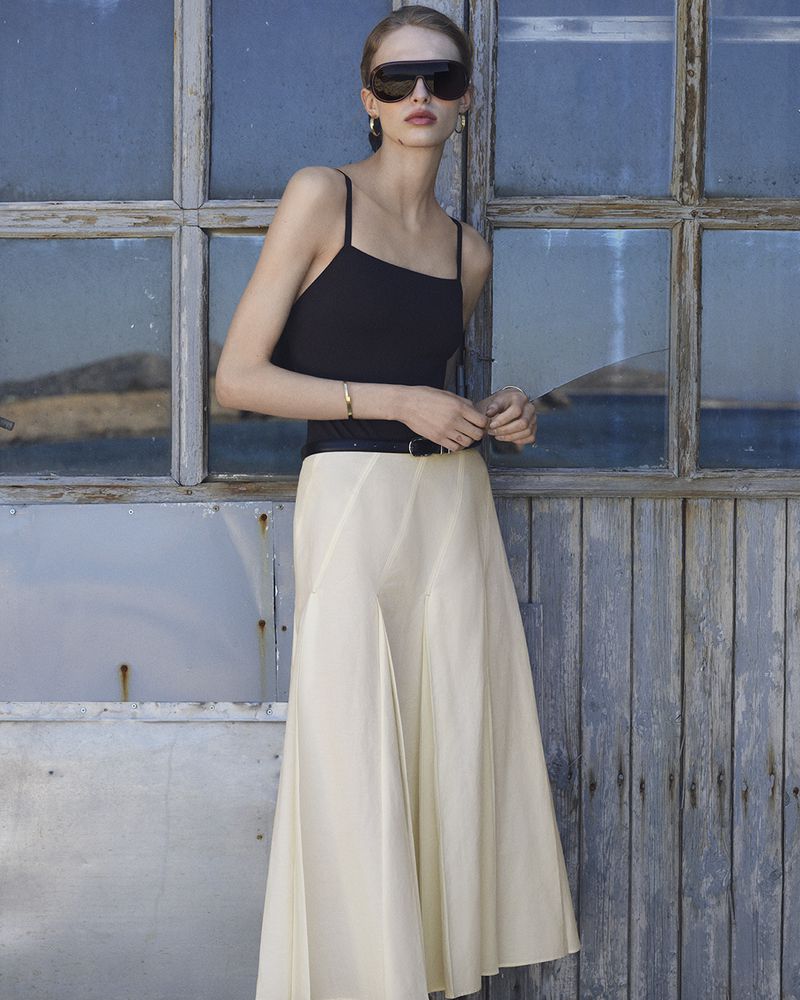 Model wearing a black camisole top, pleated cream midi skirt, and black visor sunglasses against a weathered wood and glass wall.