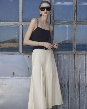 Model wearing a black camisole top, pleated cream midi skirt, and black visor sunglasses against a weathered wood and glass wall.