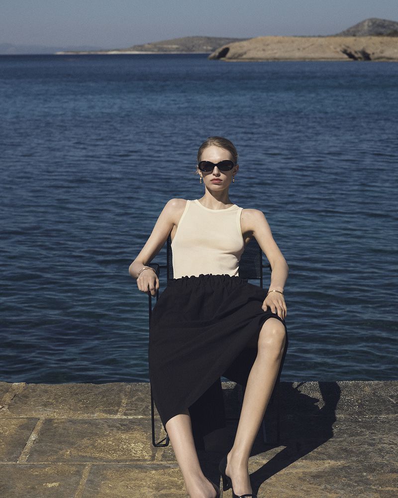 Model Karolina Spakowski wearing a cream tank top, black skirt, and sunglasses seated by the ocean for COS High Summer 2026.