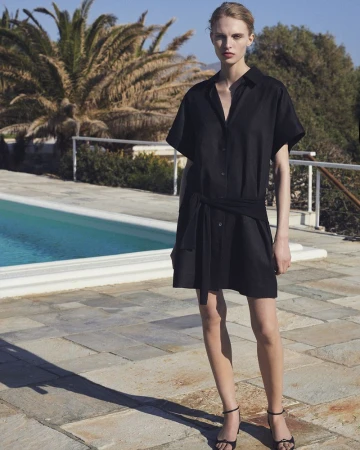 Model Karolina Spakowski in a black belted shirt dress and black strappy heels posing beside an outdoor swimming pool.