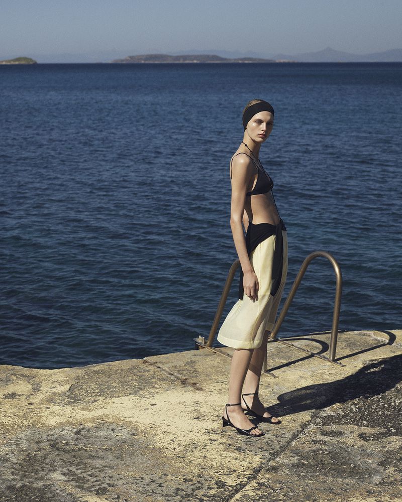 Model Karolina Spakowski in a black bikini top and pale midi skirt standing by the sea on a stone dock in bright sunlight.