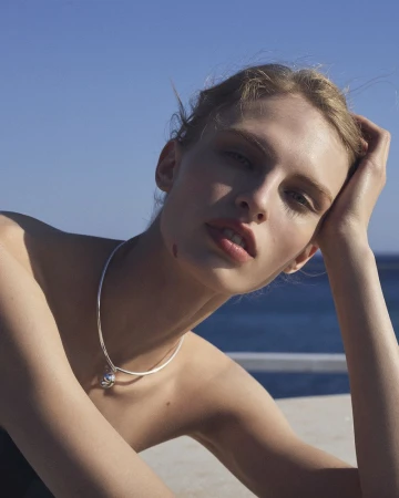 Model Karolina Spakowski wearing a silver pendant necklace and resting her head on her hand under a clear blue sky by the sea.
