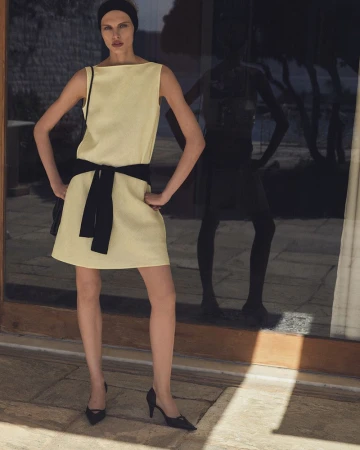 Model Karolina Spakowski wearing a sleeveless pale yellow dress with a black sash belt and black heels outside a glass door.