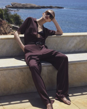 Model Karolina Spakowski in a dark burgundy jumpsuit and sunglasses reclining on a stone bench against a coastal ocean backdrop.