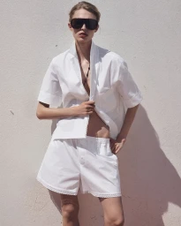 Model Karolina Spakowski wearing oversized black shield sunglasses and a white short-sleeve shirt and shorts set in bright sunlight.