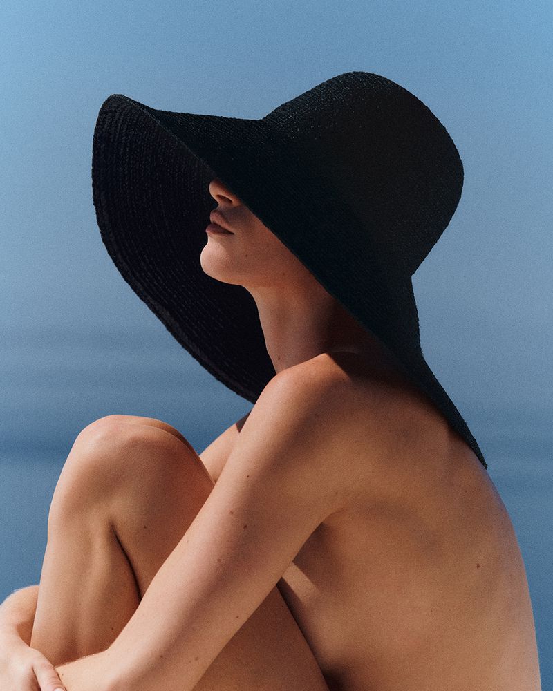Model wearing an oversized wide-brim black woven sun hat seated in profile against a clear blue ocean background.