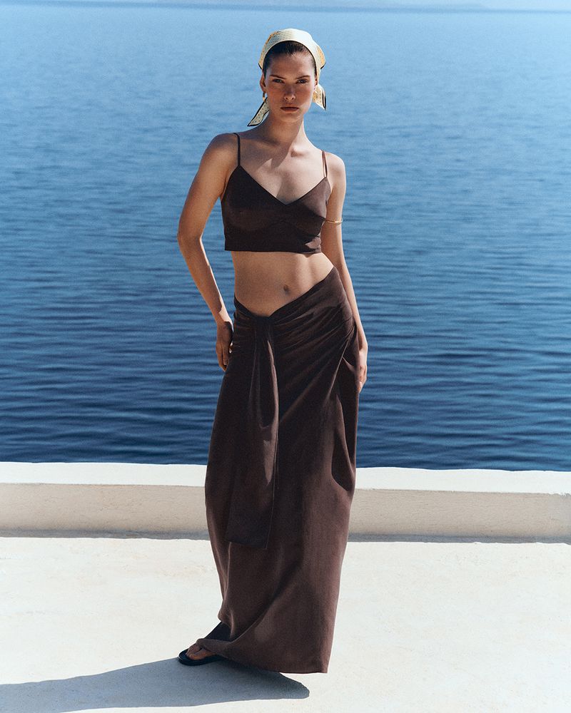 Model wearing a brown crop top, matching draped maxi skirt, and a headscarf against an ocean background.