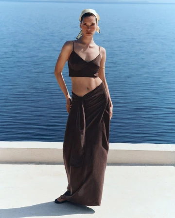 Model wearing a brown crop top, matching draped maxi skirt, and a headscarf against an ocean background.