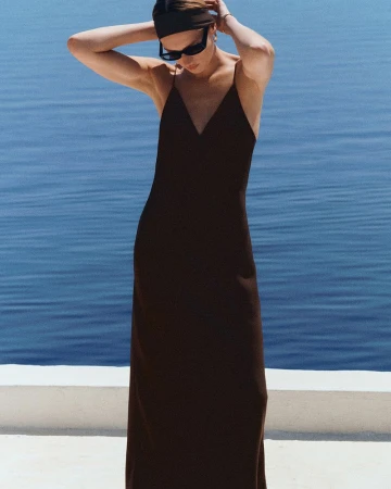 Model in dark maxi dress with sunglasses and headband standing against ocean backdrop for COS Beach 2026.