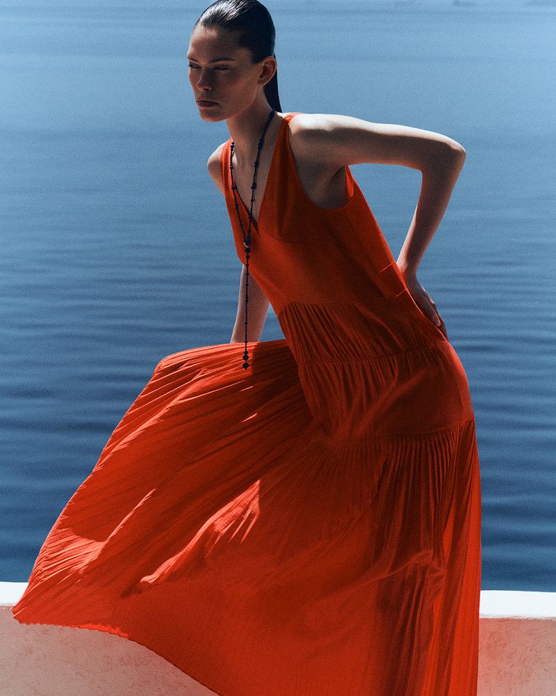 Model wearing a flowing red pleated maxi dress posing on a white ledge overlooking deep blue ocean water.