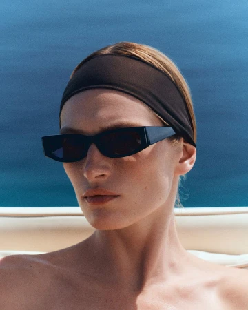 Close-up portrait of a woman wearing black geometric sunglasses and a dark brown headband against a blue ocean background.