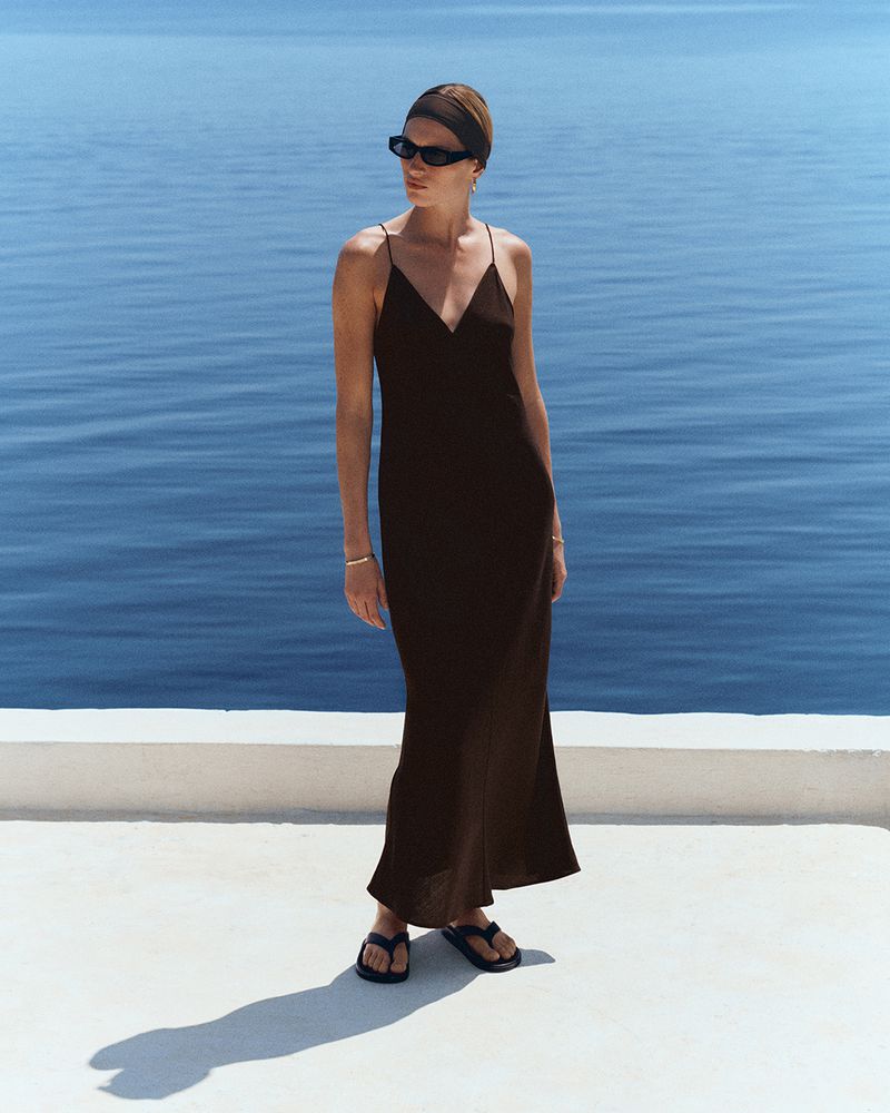 Model wearing a long black slip dress, sunglasses, and headband standing on a white terrace overlooking the ocean.