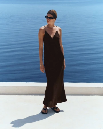 Model wearing a long black slip dress, sunglasses, and headband standing on a white terrace overlooking the ocean.