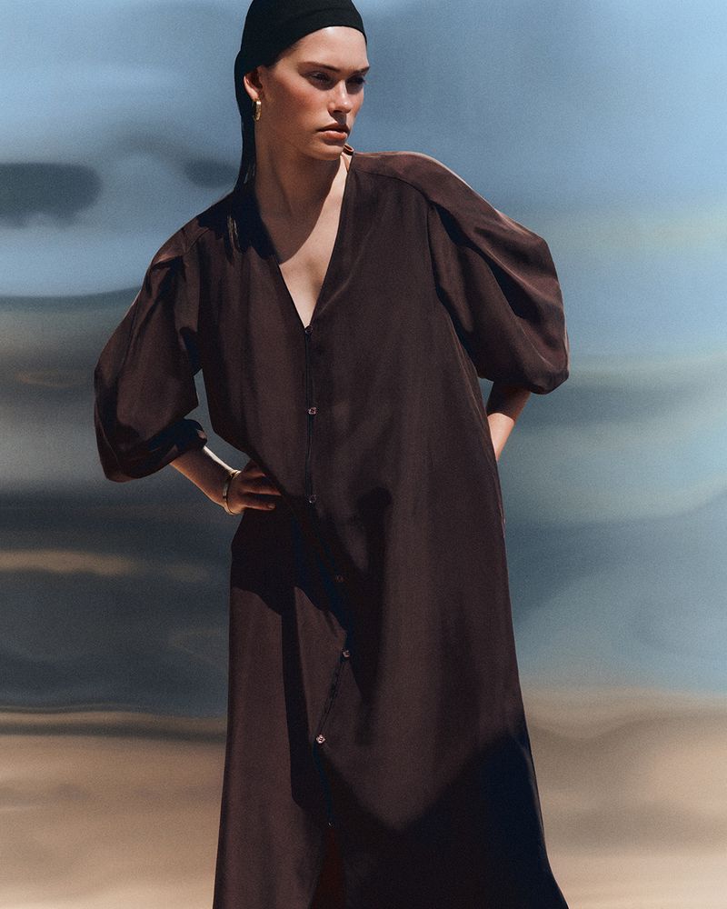 Model wearing a loose dark brown button-down dress and black headwrap posing against a blurred blue and sandy backdrop.