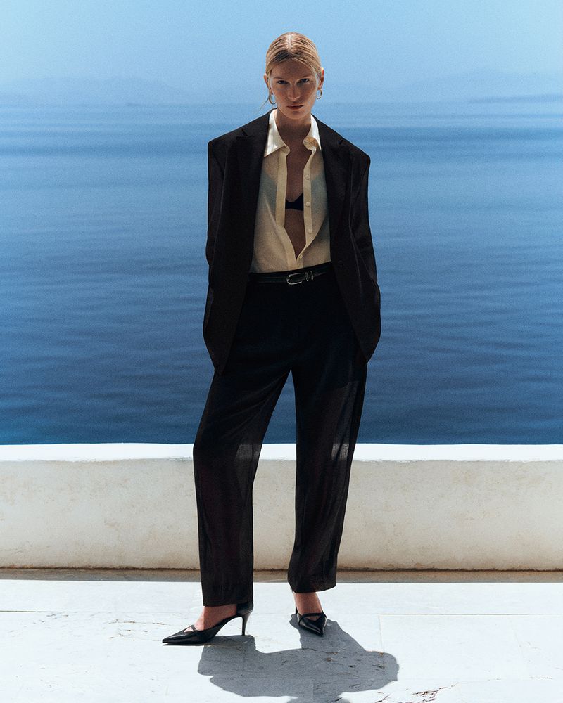 Model wearing an oversized black blazer, cream collared shirt, and black trousers against an ocean horizon.