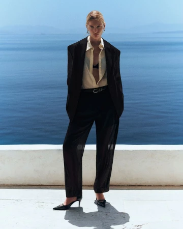 Model wearing an oversized black blazer, cream collared shirt, and black trousers against an ocean horizon.