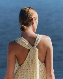 Woman seen from behind wearing a cream knotted backless halter dress against deep blue ocean water.