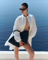Model wearing an open white tunic shirt and dark sunglasses while holding a large black woven tote bag against an open sea backdrop.
