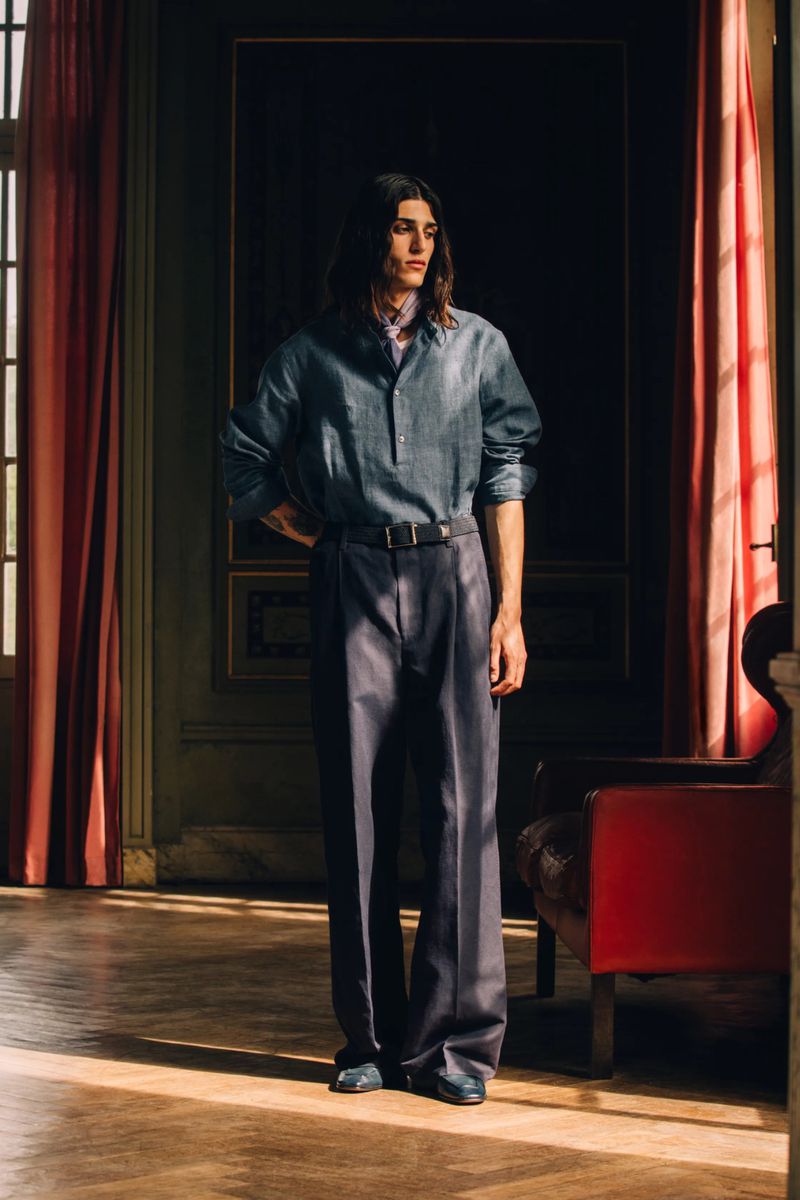 Model with long dark hair wearing a blue buttoned collared shirt and wide-leg dark trousers standing by sunlit interior curtains.