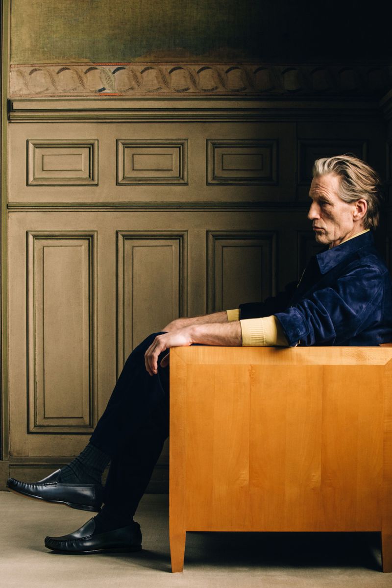 Mature man in a navy jacket and loafers seated in profile on a light wood armchair against classic wall paneling.
