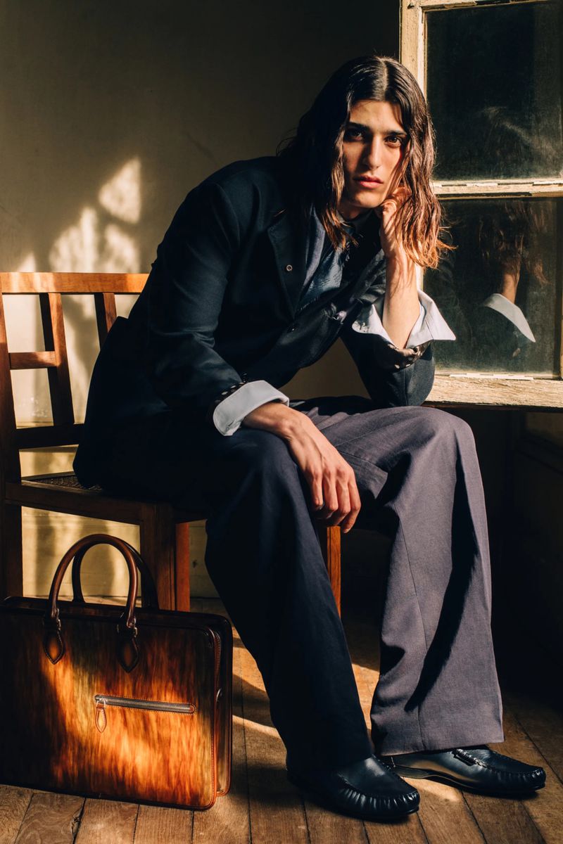 A long-haired male model in a dark tailored suit sits on a wooden chair beside a patina leather briefcase in warm sunlight.