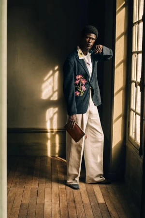 Model standing by a tall window wearing a patterned dark blazer and light trousers holding a small brown pouch