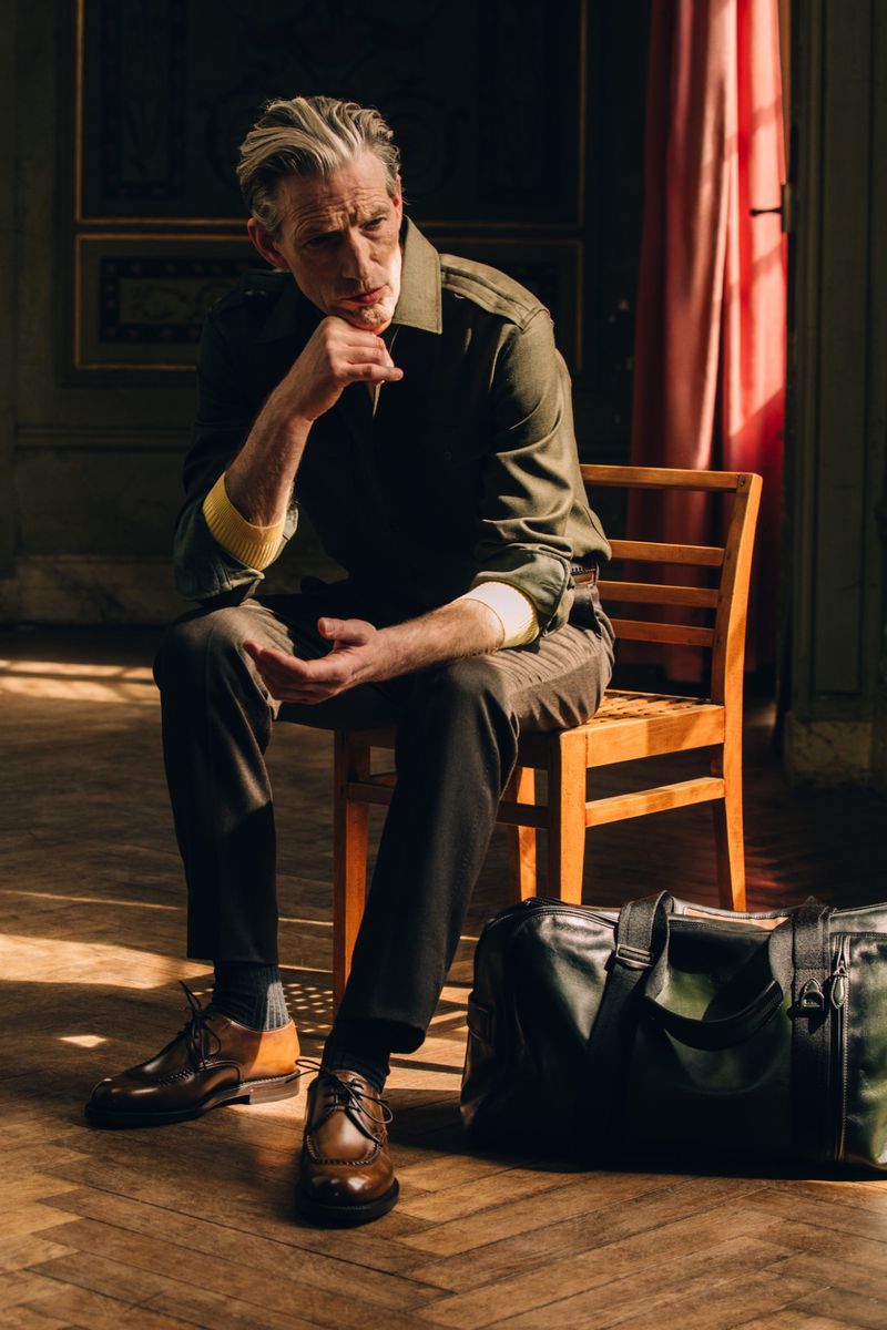 Man seated on a wooden chair wearing an olive green shirt and brown shoes next to a leather duffel bag