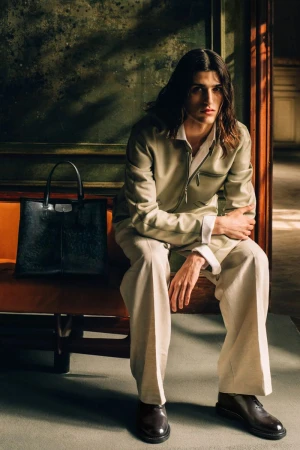 Model seated on a bench wearing a light sage zip jacket and beige wide-leg trousers beside a structured black leather tote bag.