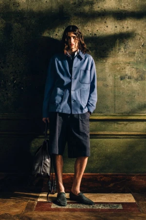 Model with long dark hair wearing a blue buttoned jacket and dark shorts standing against an aged green wall, holding a leather bag.