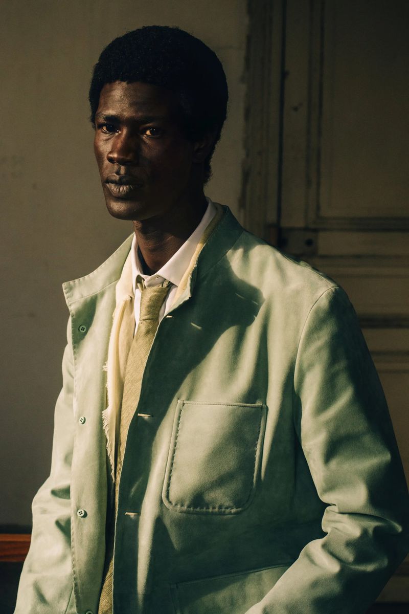 A Black man in a pale sage green workwear jacket with a collared shirt and tie, lit by direct sunlight against a paneled wall.