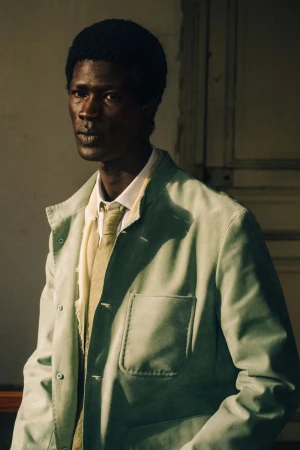 A Black man in a pale sage green workwear jacket with a collared shirt and tie, lit by direct sunlight against a paneled wall.