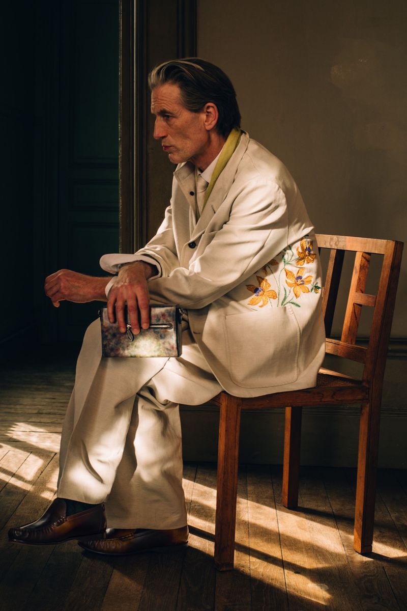 Man seated on a wooden chair wearing an off-white suit with floral embroidery and holding a clutch under directional sunlight.