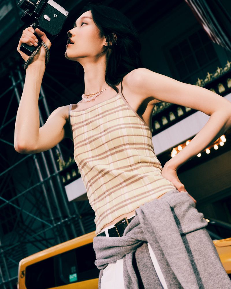 Woman in striped tank top holds vintage Super 8 camera outdoors at night with a cityscape backdrop and yellow taxi in the shot.