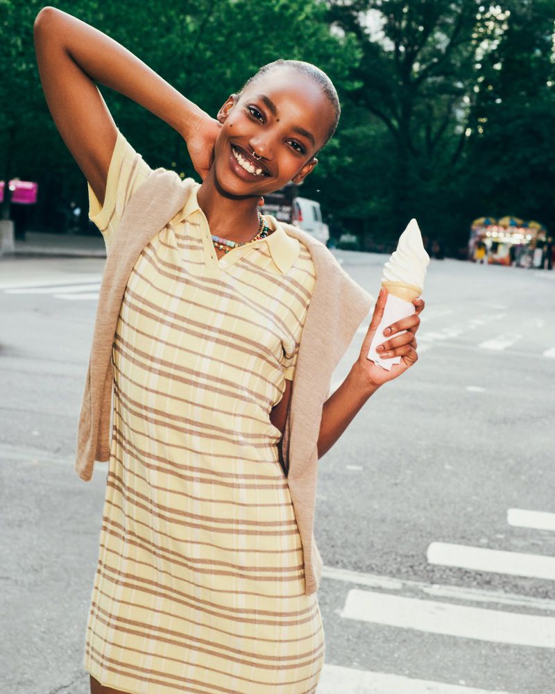 Smiling woman in striped yellow dress stands on crosswalk holding ice cream cone in city park on a sunny day, looking happy.