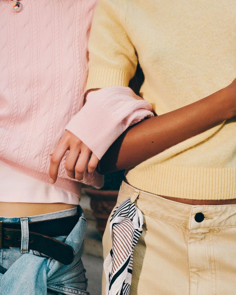 Close-up of two women in pastel sweaters and blue jeans; one wears beige pants with a scarf, shot indoors with soft lighting.
