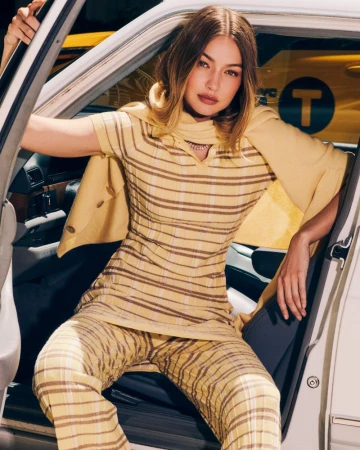 Gigi Hadid poses in a taxi cab wearing a yellow patterned top and pants. The image has bright lighting and a medium shot.