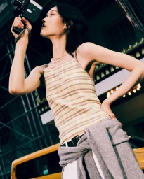 Model Chloe Oh holding a vintage movie camera outdoors wearing a striped knit tank top and grey sweater for Guest In Residence.