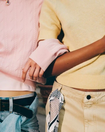 Close-up of two models linking arms, wearing a light pink cable-knit sweater with jeans and a pastel yellow knit top with a beige skirt.