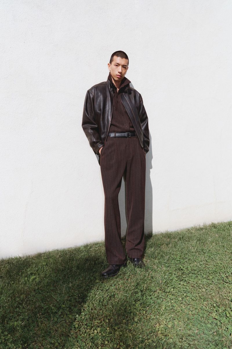 Model Cai Yuyang wearing a dark brown leather jacket and matching pleated trousers standing against a white wall on grass.